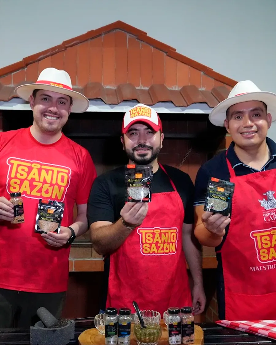 Los tres fundadores de Tsanto Sazón con delantales y camisetas rojas, mostrando sus sazonadores frente a una parrilla